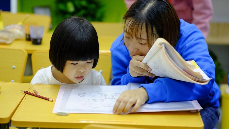 A female teacher reading out a book to a young girl as she looks at the book