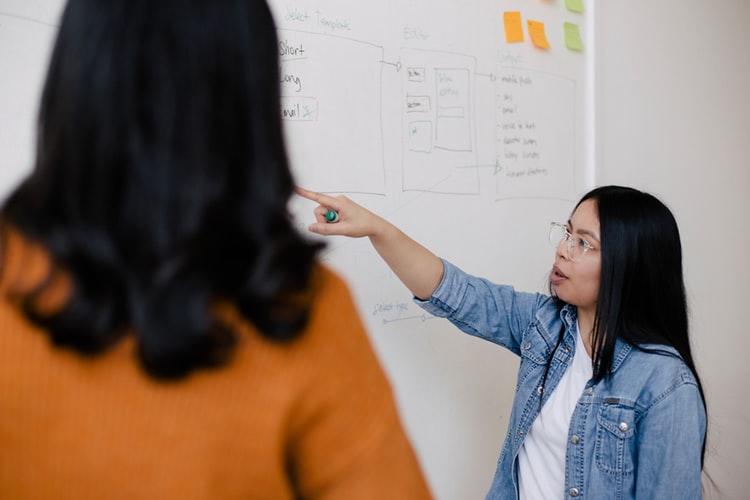 a woman points out something on a white board to another woman who is standing with her back facing the camera