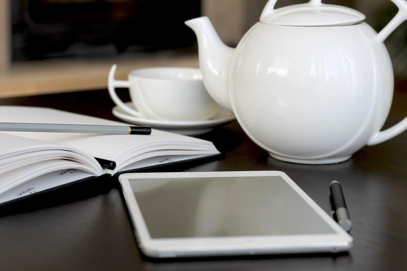 desk with a notebook, tea cup, kettle and ipad laying on top
