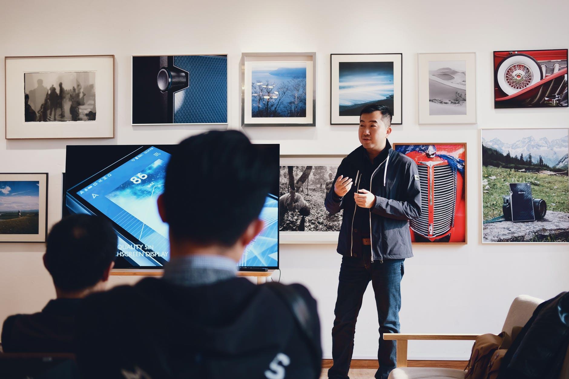 A man explains something to a class of people in front of a television.