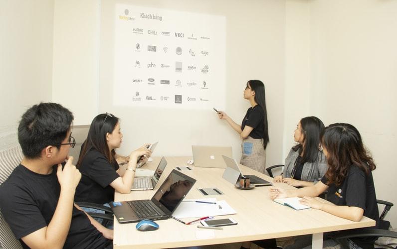 a woman pointing towards a chart on the wall as a group of seating individuals look on