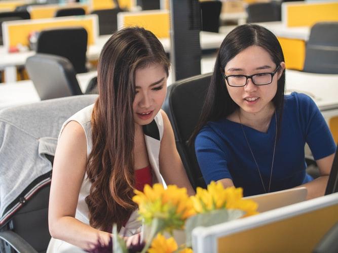 A woman helps another lady with her work in front of a computer