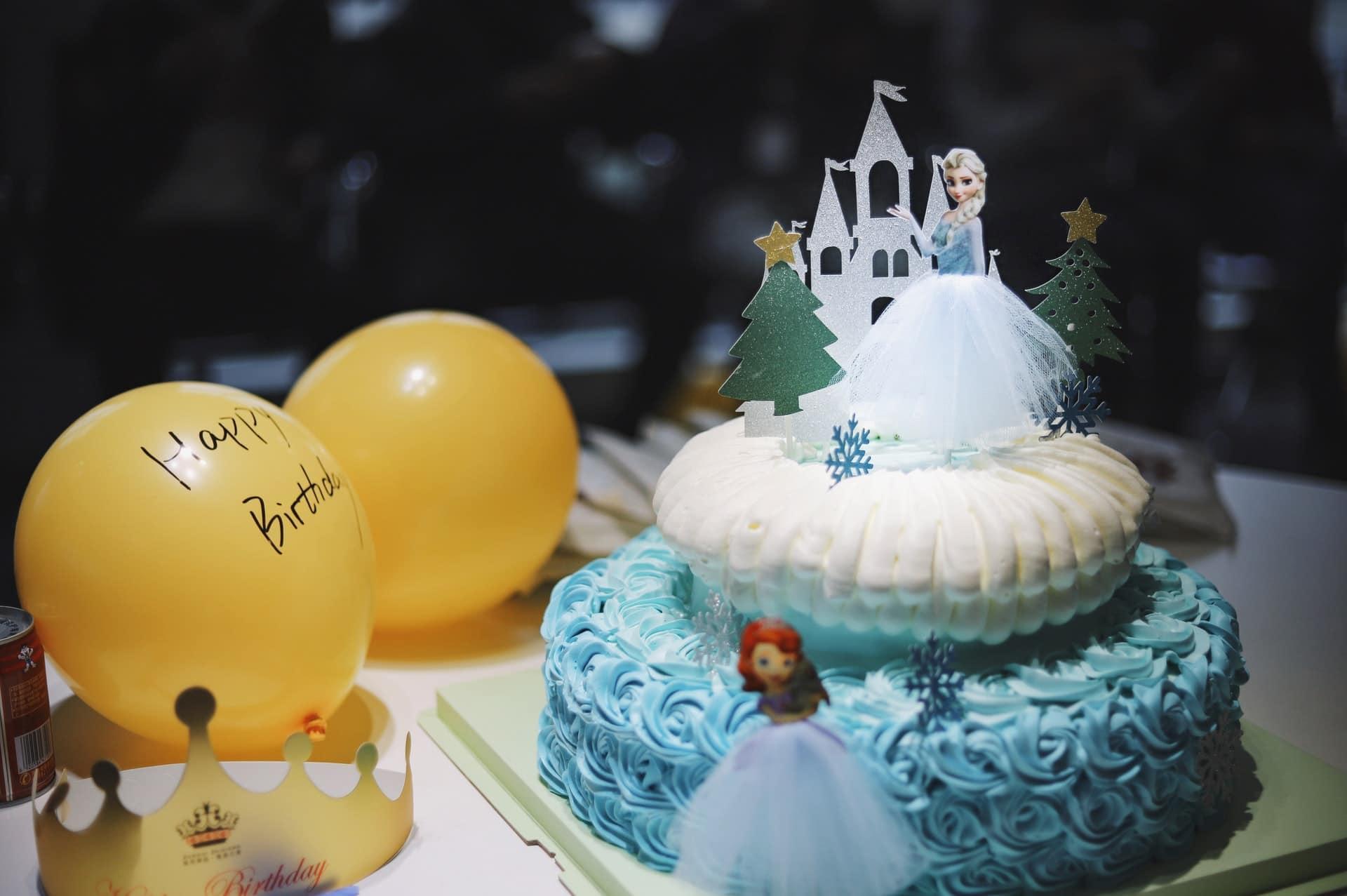 Two-layered wedding cake with princess Elsa topping, next to two yellow balloons.