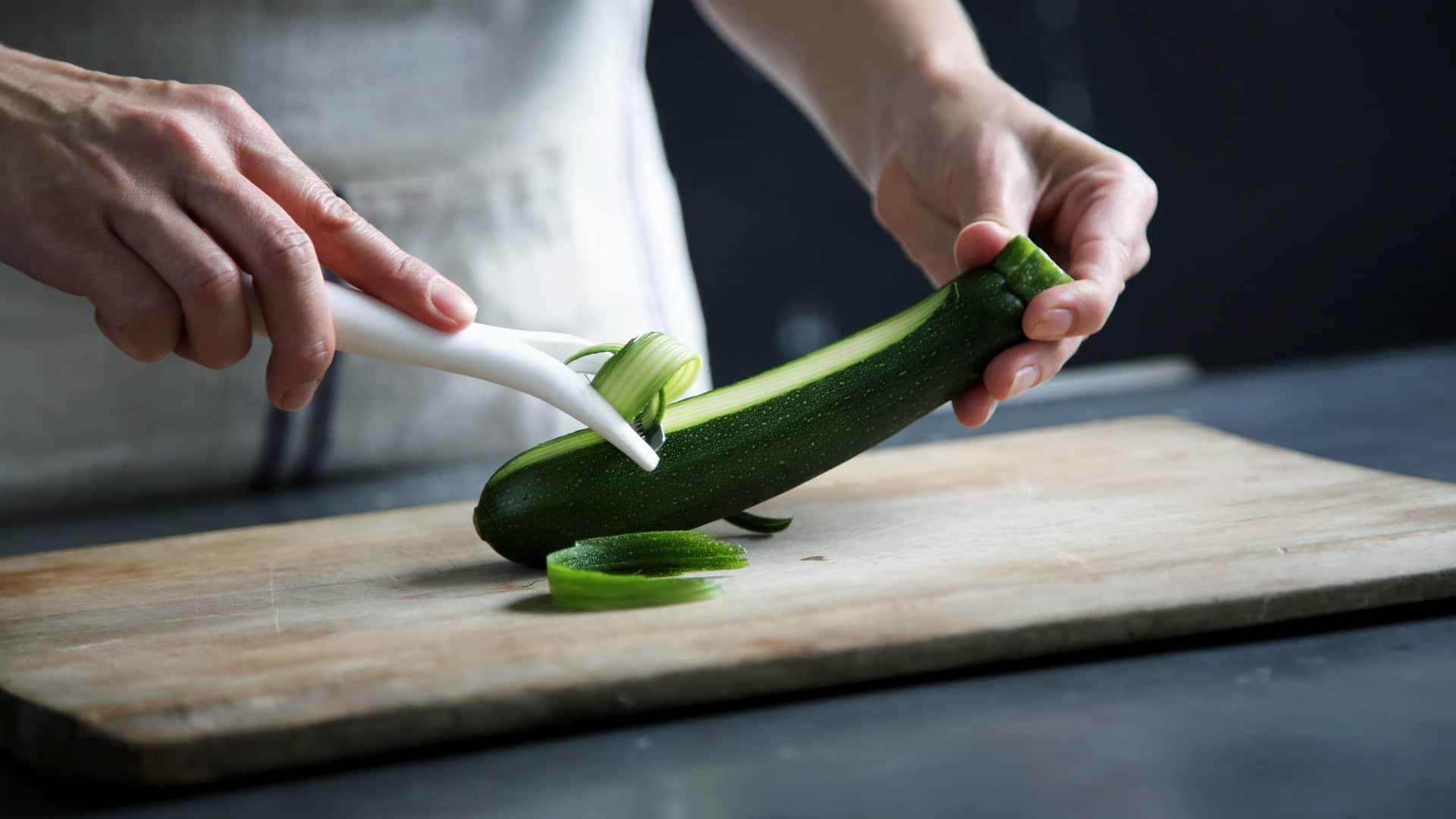 Hand peeling zucchini over a wooden cutting board.
