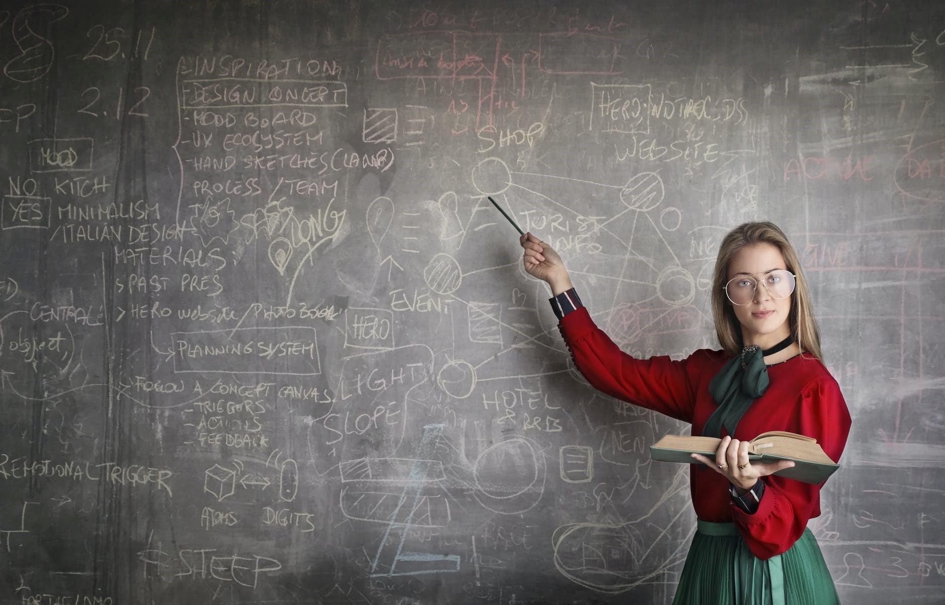 A bespectacled woman pointing towards a black board with a pen in her hand while facing the camera