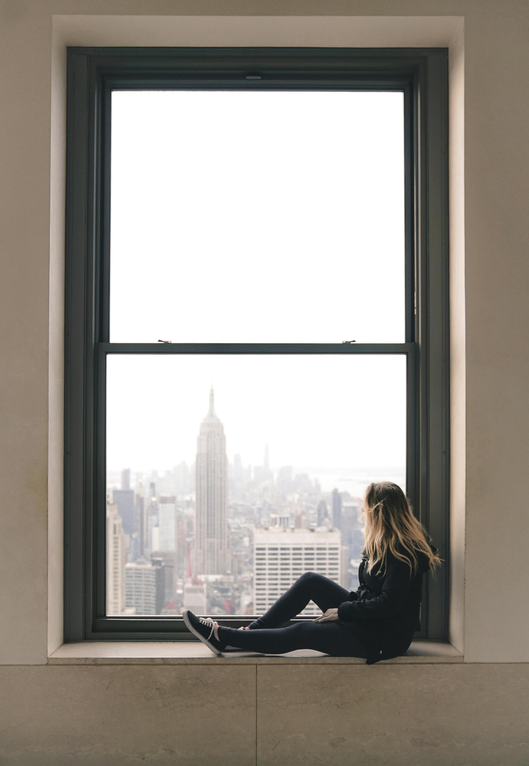 A woman sits on a window sill overlooking the New York City skyline from the Top of the Rock, gazing out at the expansive urban landscape.