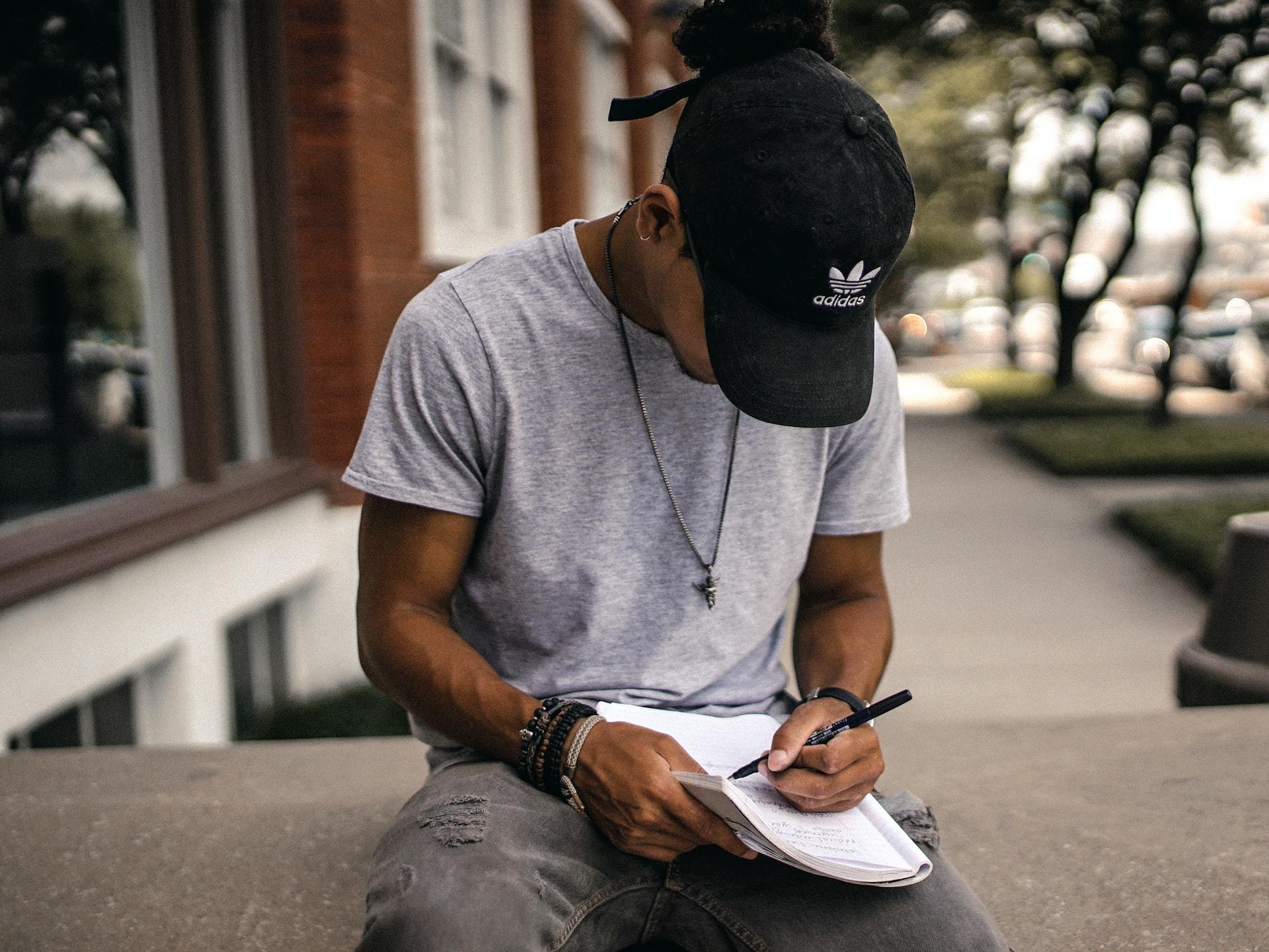 Man sitting outside on the concrete writing in a notebook with a pencil