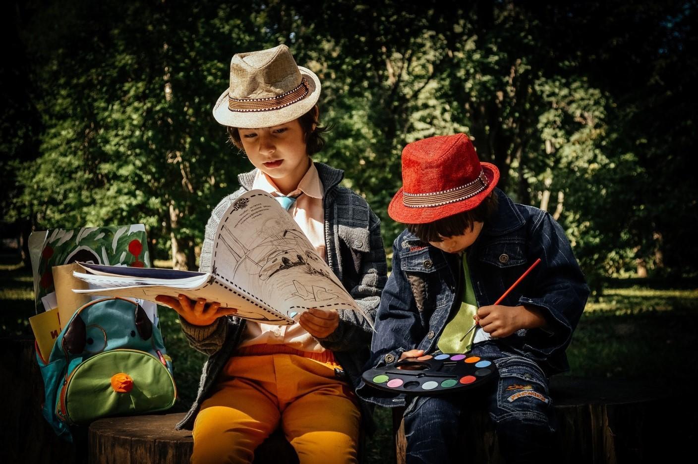 Two kids sitting on a bench, one holding a brush and a palette and the other holding a drawing book