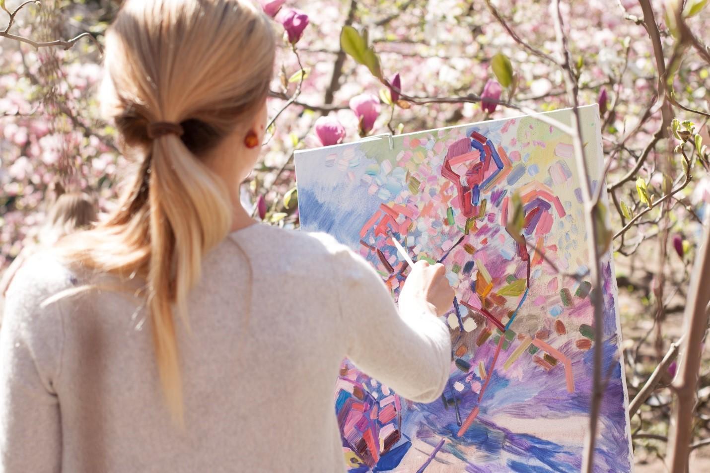 A girl is painting the autumn tree before her with a paintbrush on the canvas.