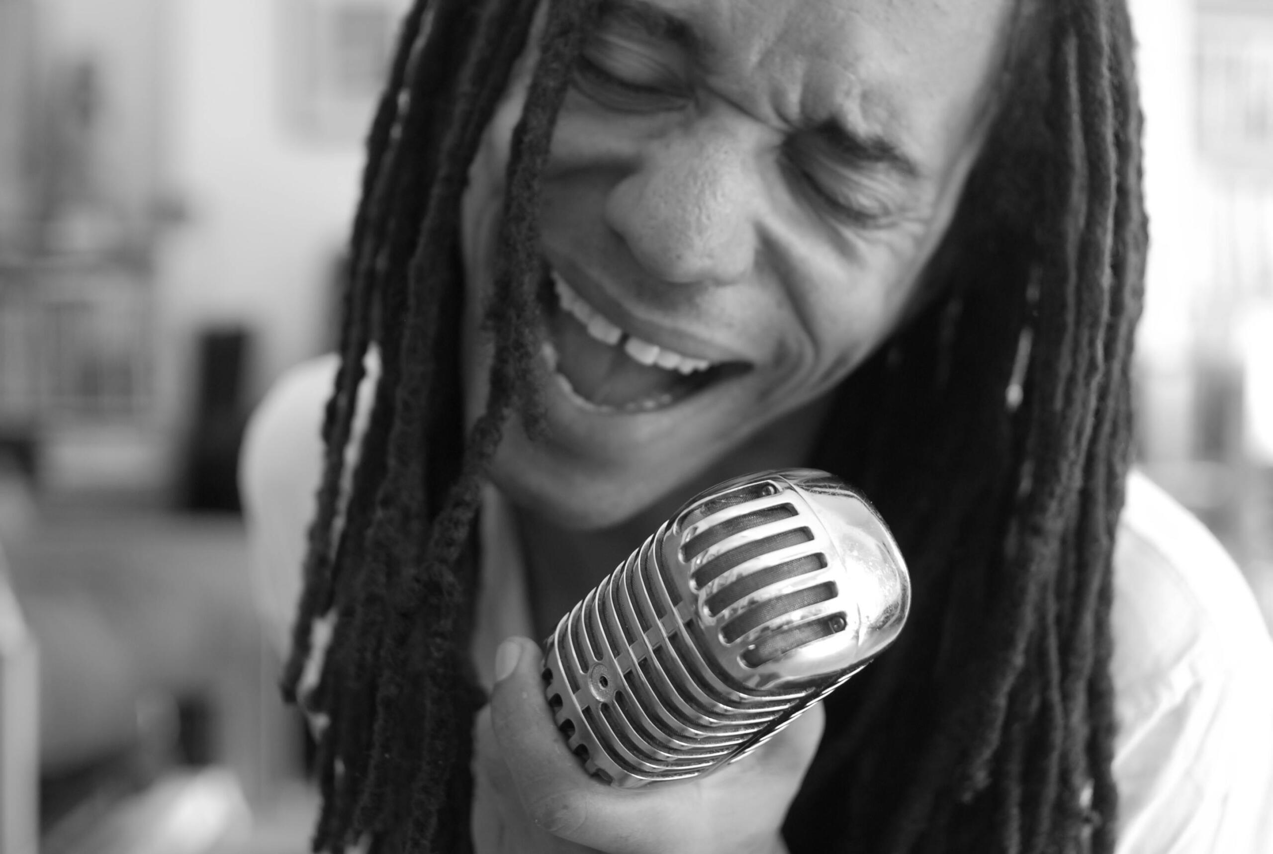 A man with dreadlocks passionately singing into a microphone in a dimly lit performance space.