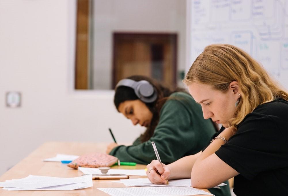 Two girls are seen sitting and solving an exam; one has headphones on her head. There is a pouch, a cellphone, and papers on the desk as well. Group study sessions provide an interactive way for students to study math; however, nothing is better than hiring a private math tutor to help you air out your inefficiencies