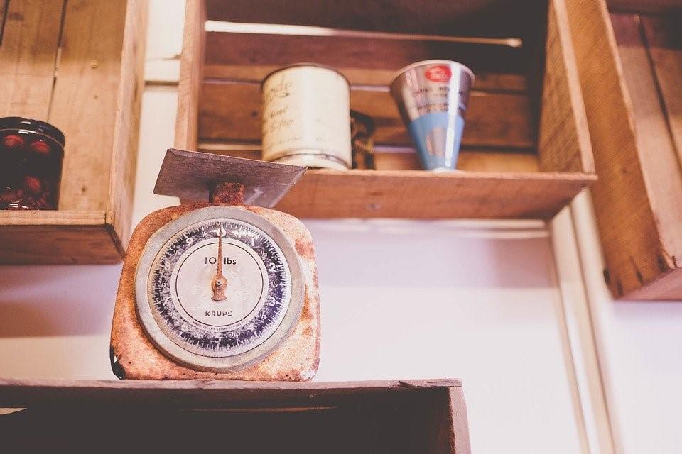A kitchen scale is present on the counter; however, it is quite an old-looking one. There are also different containers on the shelves in the background with a conical blue-colored container. Finding a math tutor will help you understand the importance of new and old measuring instruments