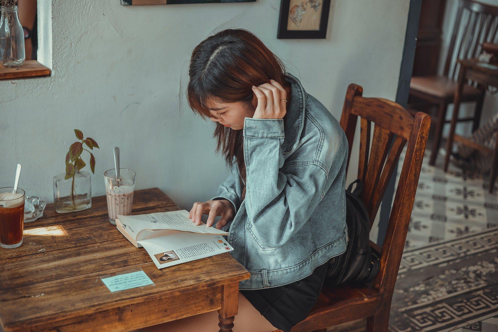 A woman wearing a denim jacket sitting on a wooden chair and reading a book. It doesn't matter what your profession or lifestyle is; you have probably tried to read as a hobby at some point in your life!