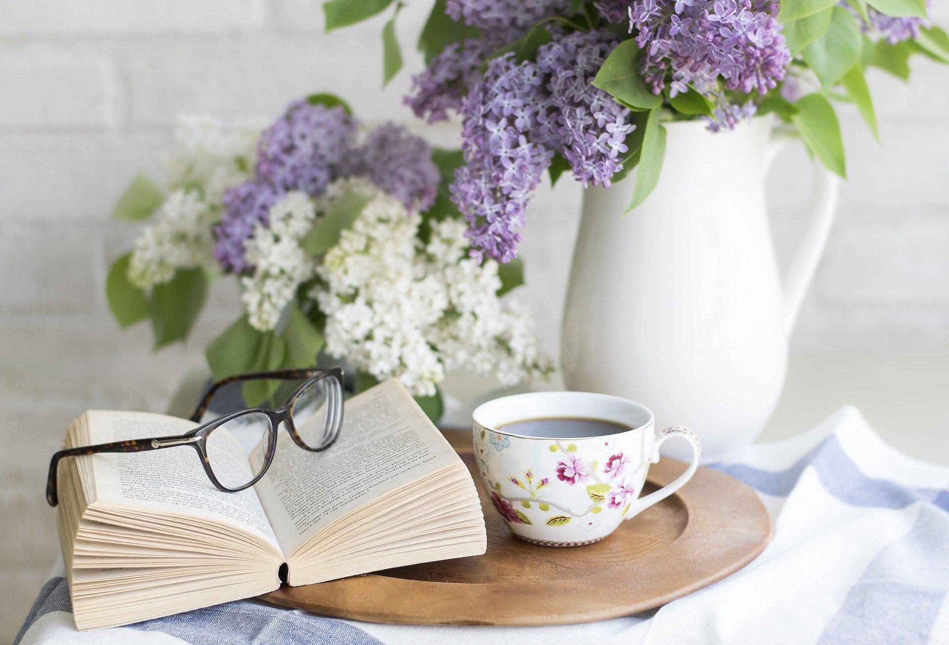 An open book, a pair of glasses, and a cup full of tea placed on a wooden tray with a white vase and flowers in the background. Hobby reading is a pleasant experience, often a therapeutic end-of-the-day activity