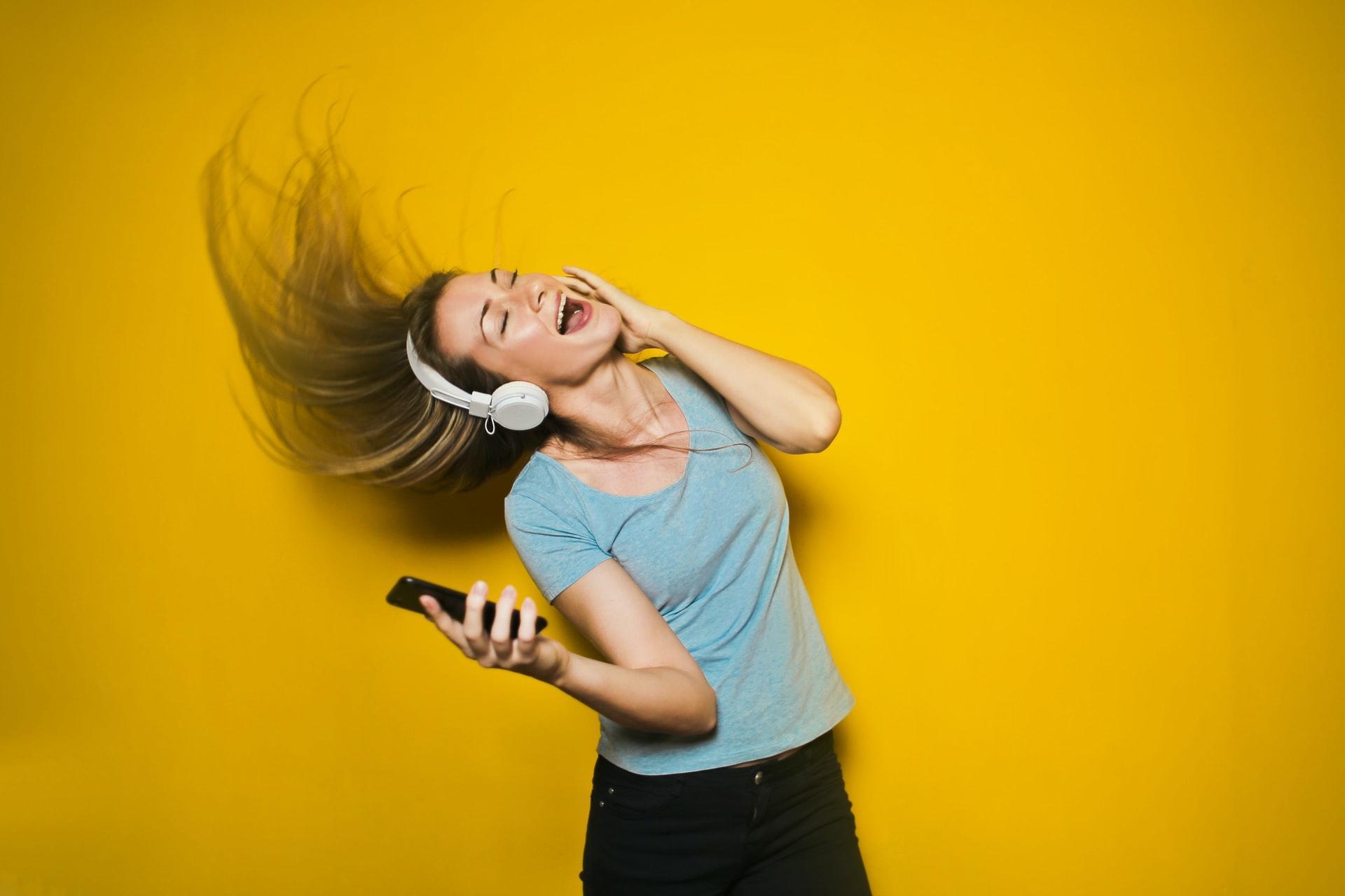 A girl swinging her hair and singing with headphones standing in front of bright yellow background.