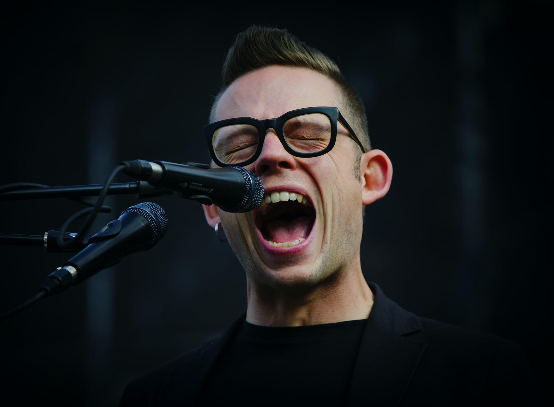 Closeup of man with glasses opening his mouth wide to sing in front of microphone.