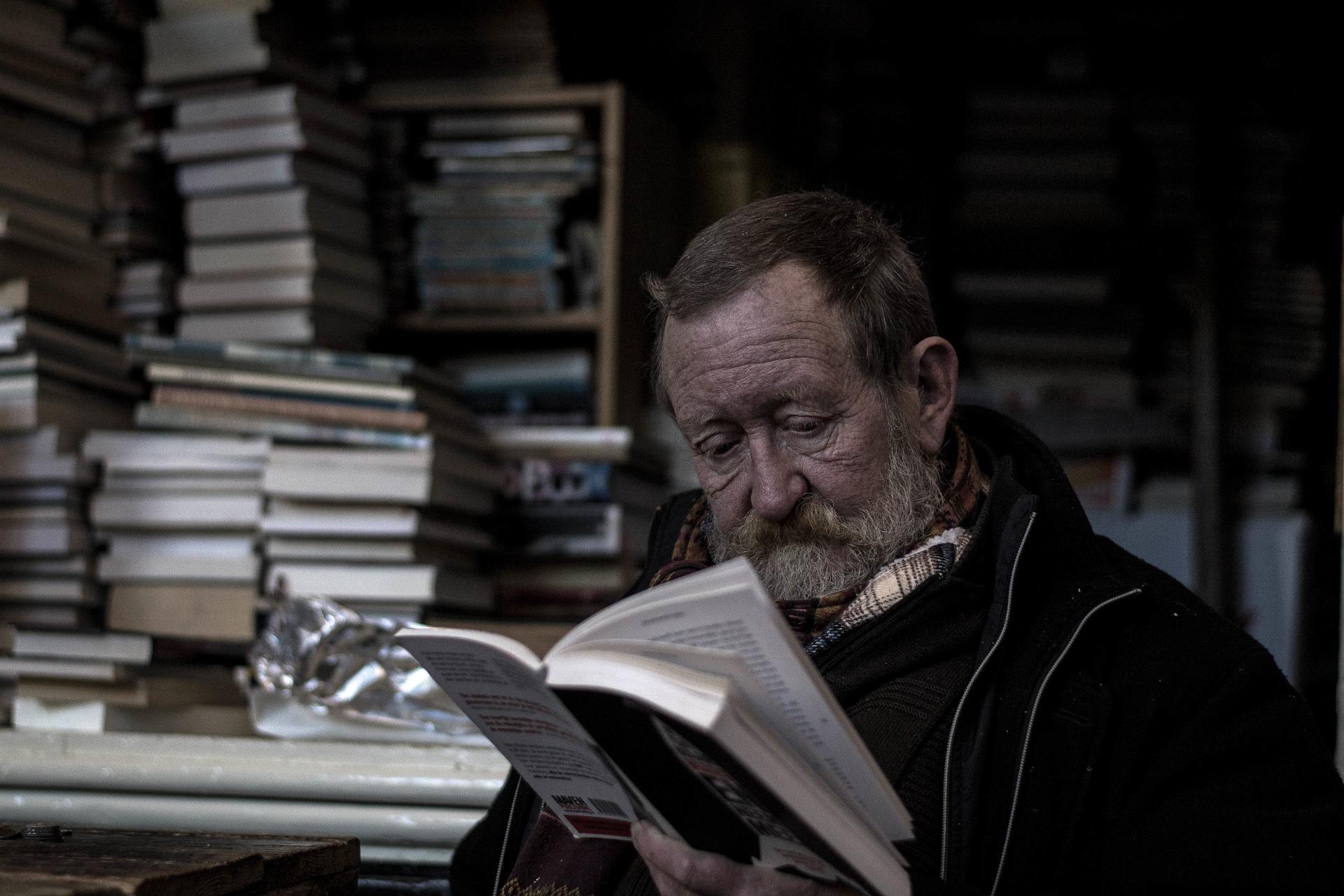 An older man with a white beard reading a book while a pile of books is stacked behind him in the dark background. Reading knows no age; while it is true that language retention is better at a younger age, critical skills are at their peak in old age