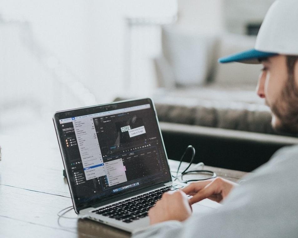 A man in a white shirt and baseball cap working on a laptop set on a wooden table. Working from home requires you to multi-task; however, the low-pressure environment helps make it easy to tackle