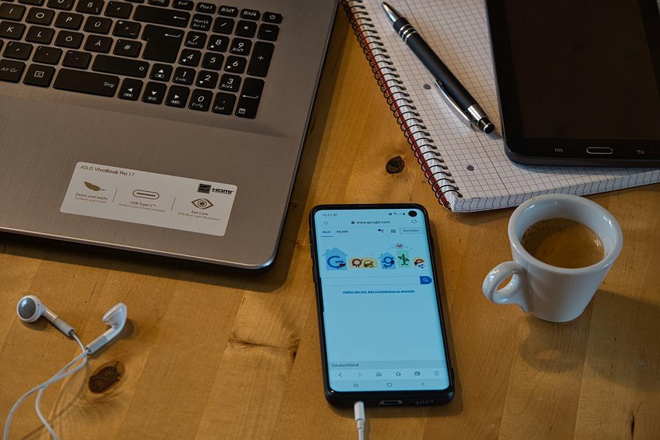 A laptop, tablet, smartphone, earphones, notebook, and pen spread out on a wooden table. A cluttered workstation mirrors a cluttered thought-process; when working from home, try to develop some discipline as it will only help you perform better