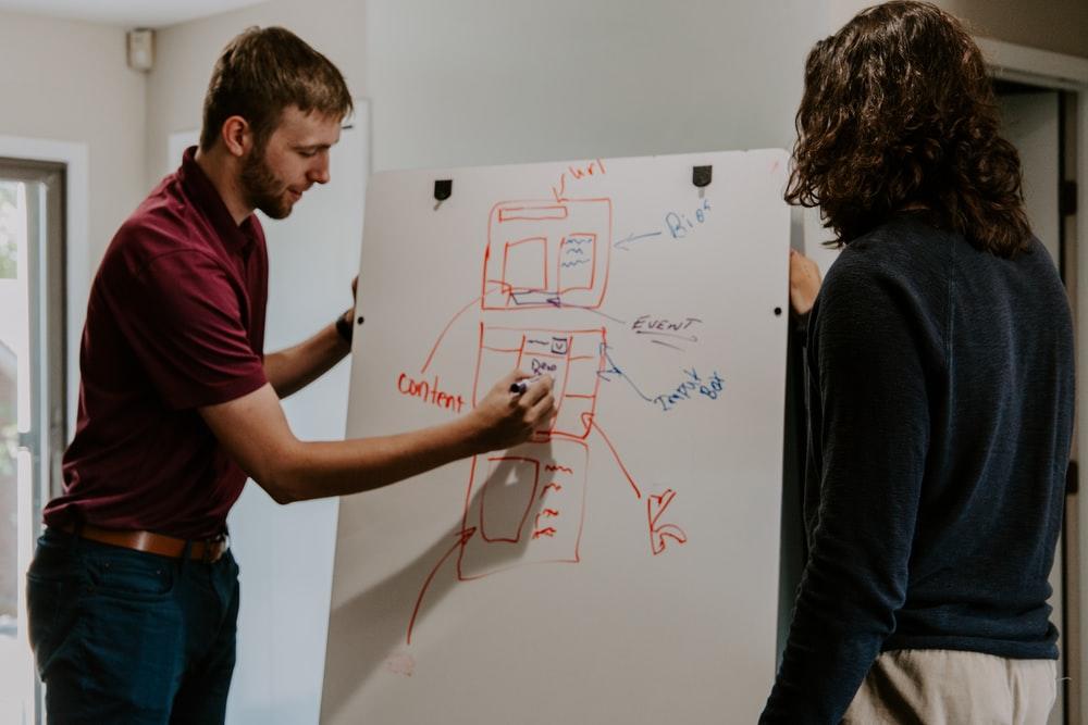 A man in a red shirt drawing a diagram on a whiteboard with another person in a blue shirt standing by his side. Private tuitions are a great way to make some money if you like working from home; teach online to as many students as you can