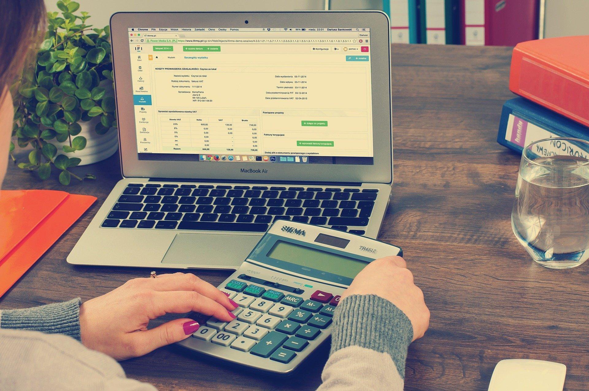A laptop, calculator, glass of water, and binders on the table. A woman is using the calculator while the accounts are open on the computer. With new work-from-home guidelines, many professionals have suffered; however, accounting jobs are as in-demand as they always were!