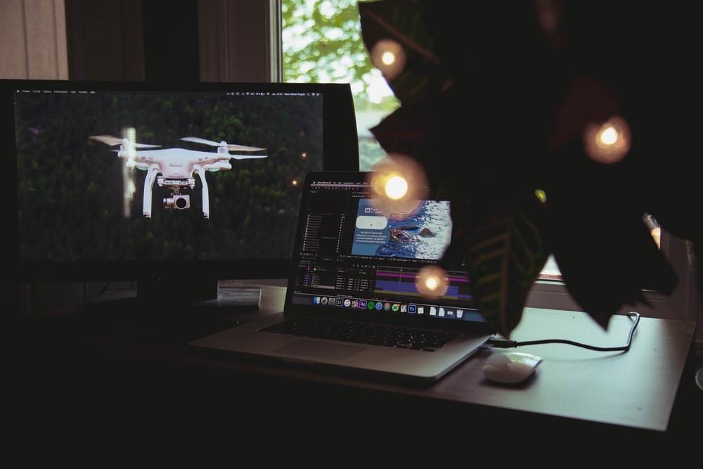 There are a desktop, mouse, and laptop on the table. The laptop is partially hidden due to the leaves. A drone picture is visible on the desktop computer, whereas Adobe After Effects is open on the laptop