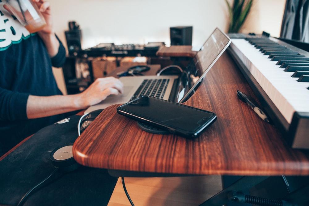 A man working on a laptop placed on a wooden table with a keyboard lying in the background. Online Adobe After Effects is a good option for students who don't have the time to commute to classes. It's a great way to learn while you work on side projects from your home