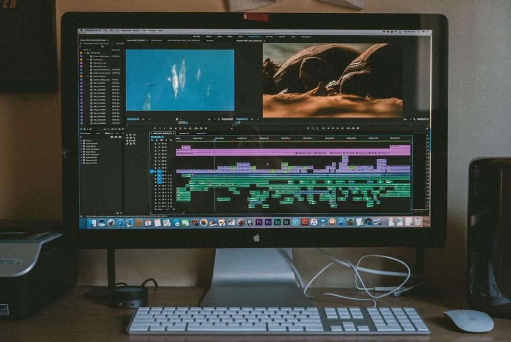 Adobe After Effects open on iMac, a keyboard and mouse are on the desk. On the left-hand side, there is also a printer present. Some of the best documentaries we see on TV, even those narrated by David Attenborough, have been enhanced using AE!