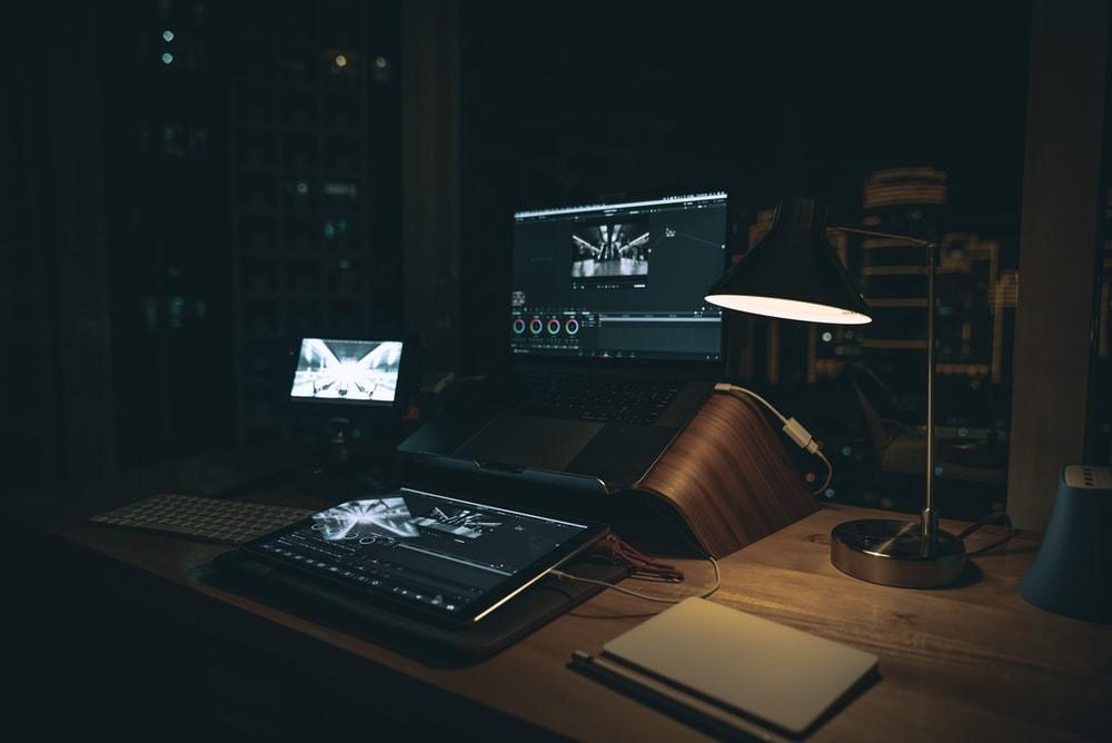 A laptop, graphic tab, lamp, a screen, and a keyboard are present on a specialized table for work. Behind the laptop, the night scenery and building can be observed. Adobe after effects works with cloud capabilities, meaning that you can shift between devices and still pick up from where you left off