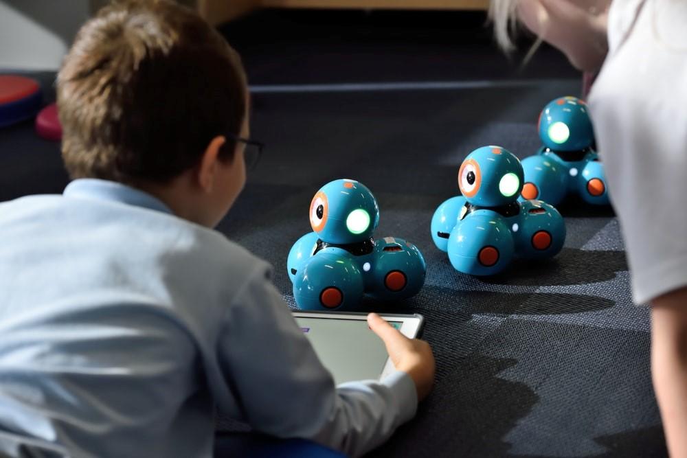 A kid using a tablet and playing with programming for kids toys and tools. There are three programming toys on the ground. An adult is overseeing the kid playing