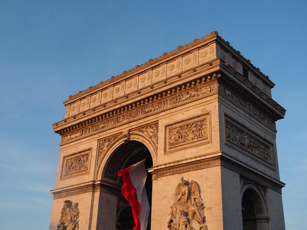 A picture of the Arc de Triomphe with the French flag hanging from the arch taken during the daytime. France is an architectural enthusiast's paradise, offering some of the most notable landmarks of European architecture