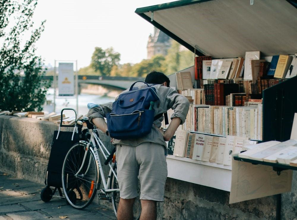 A man wearing a backpack while browsing through a roadside bookstall. The bookstall is filled with French books, and there is also a bicycle beside the book stand. The books are also lined up on the stone hedge, and a river can be seen in the background