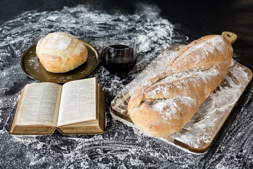 A bread on a chopping board, an old book, another loaf of bread on a copper plate, and a glass of wine. There is also flour spread all over the table. French language lessons will help you read authentic recipe books and have your go at French cuisine