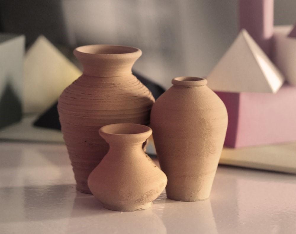 A set of three clay vases with a natural brown tone resting on a white surface. Some of the most basic pieces you can make in your pottery lessons include household utensils like bowls, mugs, and vases