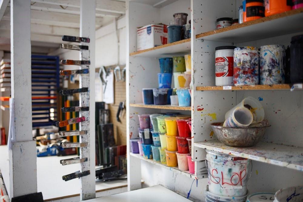 An inside view of the art supply closet. There are multiple paint buckets lined over the shelves, as well as other art supplies. Numerous clips are clipped on a single wooden frame. An art school will usually have several messy studios and experimental paint rooms for you to work in