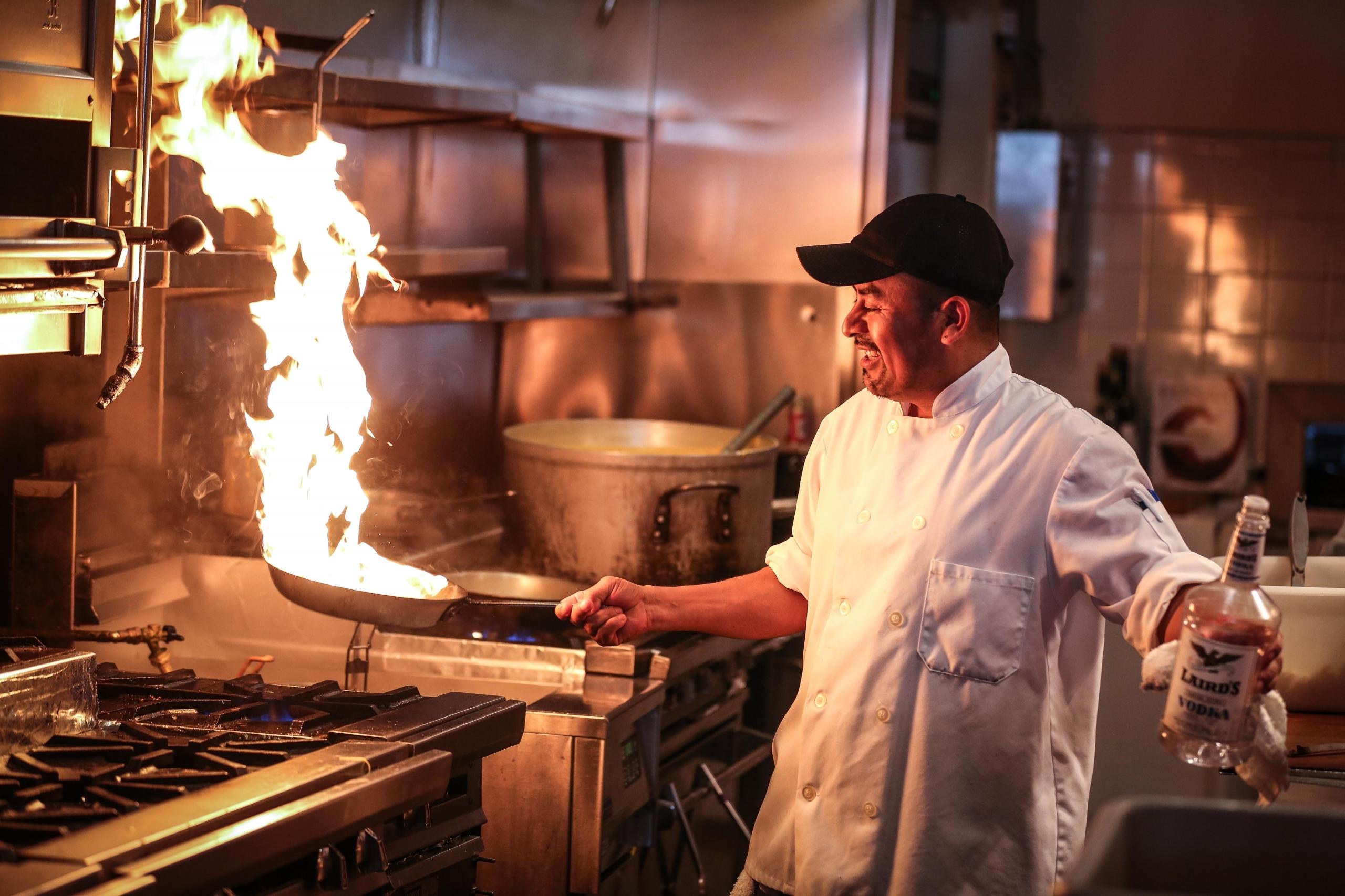 A chef smiling as he watches flames erupt on the frying pan after he pours vodka. The chef is wearing the traditional white coat and holding a frying pan in one hand and a vodka in the other. If you want to become a chef as revered as Anthony Bourdain, you must love what you do
