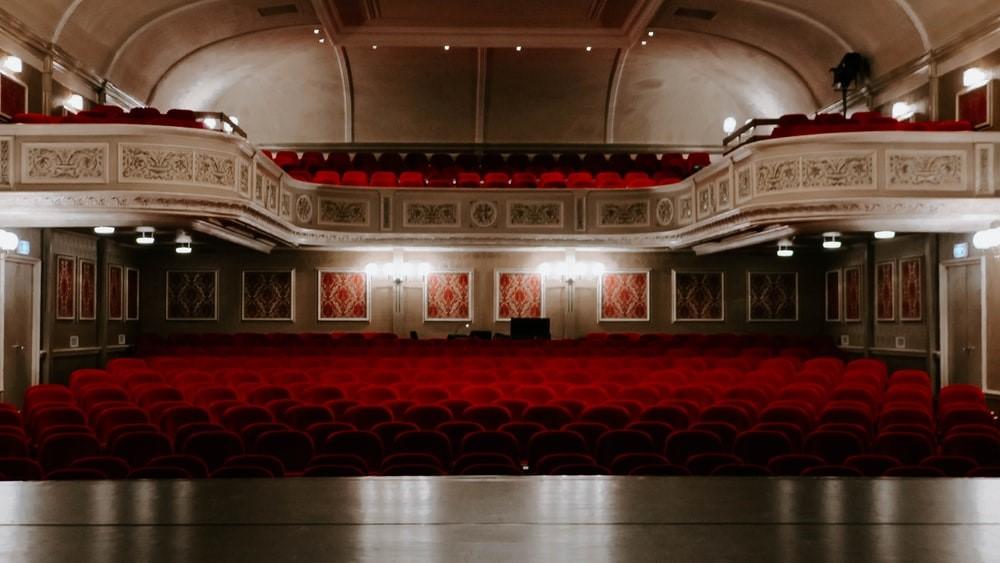 An empty multi-tiered theater room with dim lights and red seats as seen from the stage. The Palace is one of the most famous theaters in Broadway, New York, known for its high-end shows