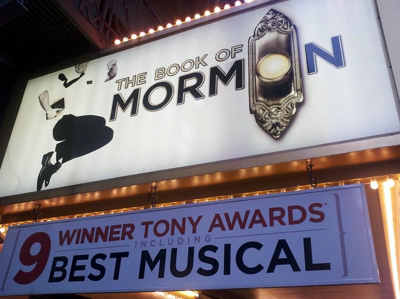 A poster of The Book of Mormon musical on Broadway, above another poster that states, "Nine-time winner Tony Awards including Best Musical." Walk down Time Square, and you will probably see Book of Mormon adverts every few steps!