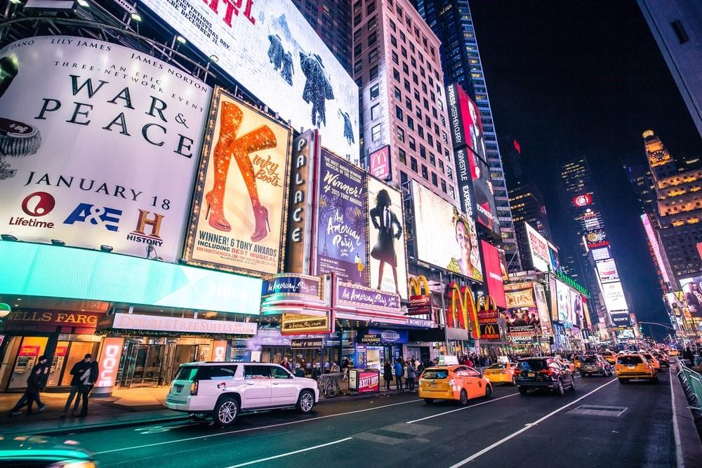 A New York City road with signboards showing the running Broadway shows at the famous Palace Theatre and the Times Square visible in the background
