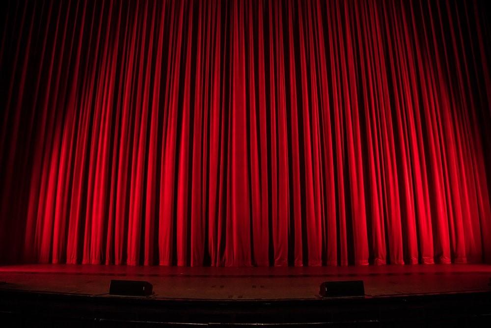 Ceiling high red theater curtains pulled on a wooden theater stage. The neighborhoods of Broadway and West End are seen as synonymous with high-end theater and house some of the best theaters in their respective countries