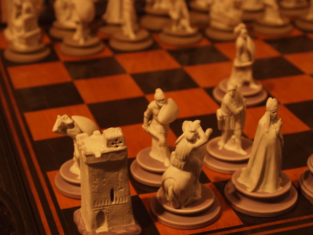 Customized chess pieces lined up on a brown and black chessboard. The rook, knight, queen, and pawns are visible. Playing chess requires competitors to verse themselves on what the pieces are called and how they move