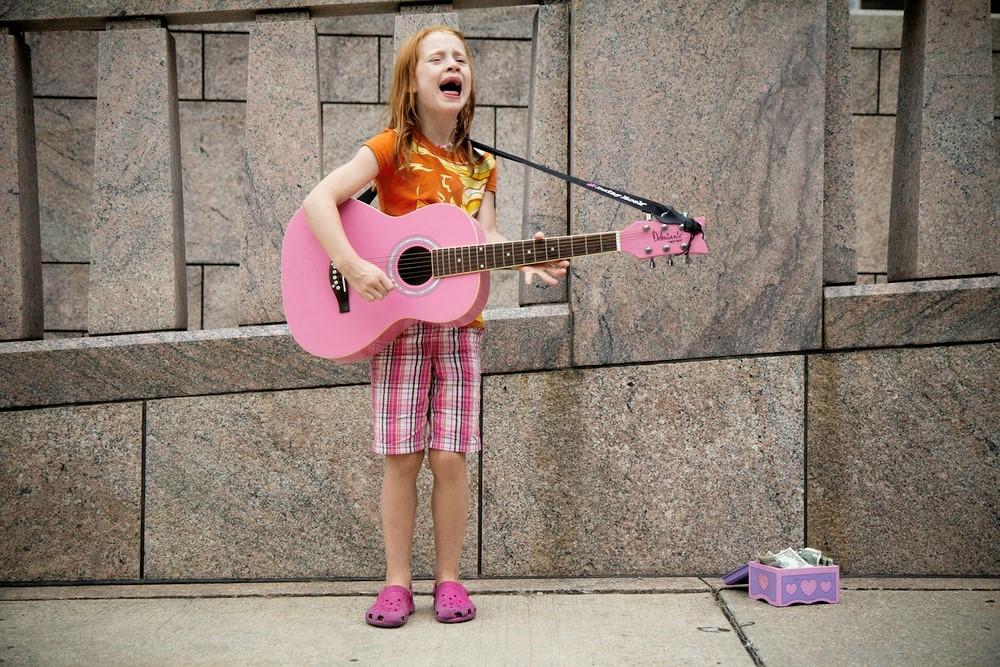 A young girl in shorts and a t-shirt singing and strumming a pink guitar with a purple money box placed by her side. Most musicians started off singing as young children. Nowadays, young children who are into singing can resort to using a voice training app