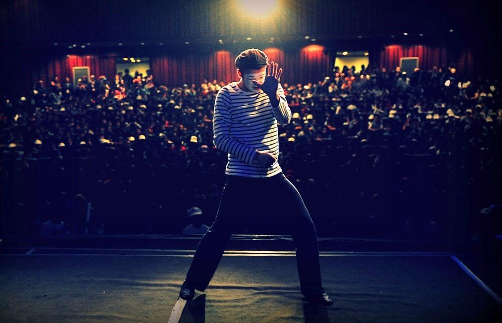A pantomime artist is wearing a white striped shirt and beret, miming with his back towards the audience. To learn more about theater and how famous Broadway shows work, sign up with a trained acting coach on Superprof