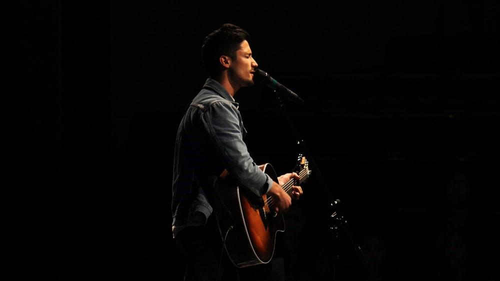 A man wearing a denim shirt holds a brown guitar and sings into a microphone in a dark setting. Training your voice to avoid injuries to your vocal cords and throat is monumental for professional singers