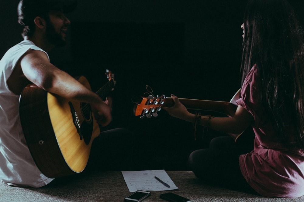 A guy and girl sitting in front of each other holding guitars and singing. There is a piece of paper on the ground and two cell phones. In case you find your throat hurting after a singing session, get yourself checked for a potential vocal cord injury