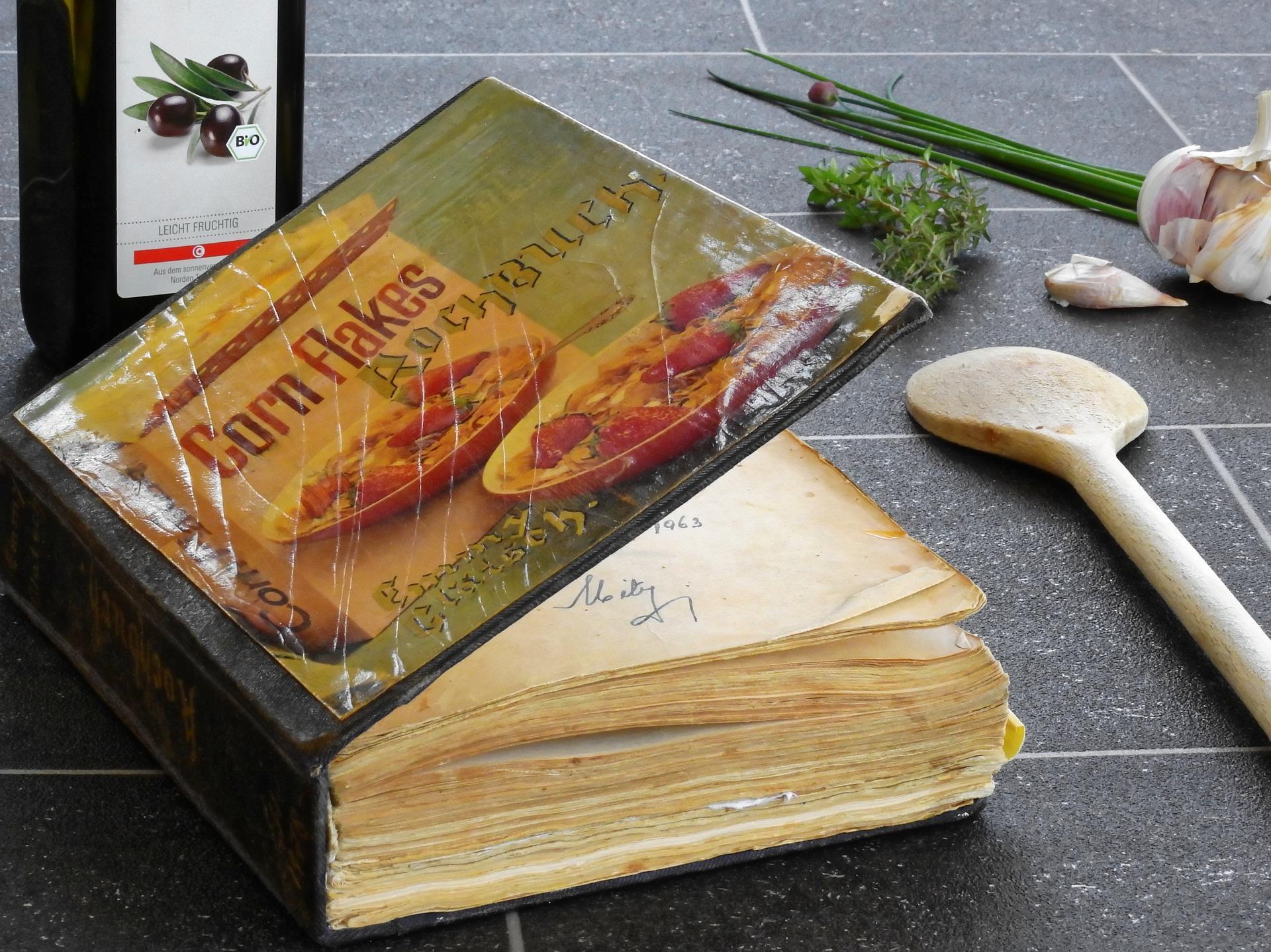 An old hard-cover cookbook on a kitchen counter. There is also a wooden spoon, oregano, garlic, and green onions next to it. Julia Child's cookbook was a sensation among the masses as it presented recipes with such intricate depth