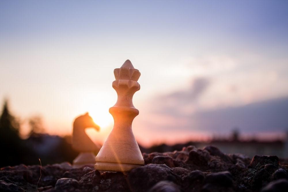 A white bishop piece and a knight piece placed on rocky ground with the sun setting in the background. Learn to play chess online without the hassle of commuting to chess classes every week with Superprof!