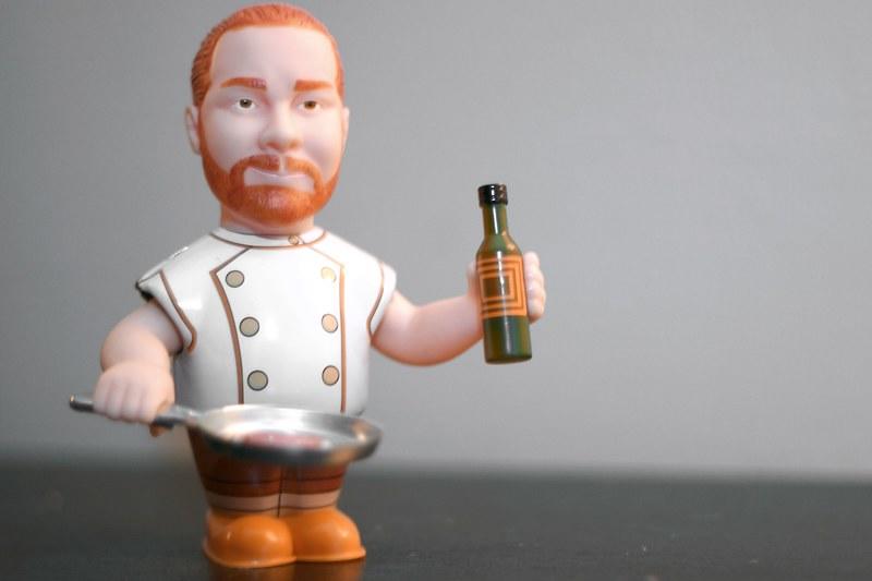 A mini bobblehead figurine of Mario Batali, the famous chef, holding a sauce bottle in one hand and a frying pan in the other. Many food chefs who become tv stars end up getting a line of dedicated merchandise and collectibles as well