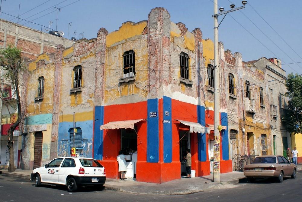 A white car is parked beside a decaying building colored red, yellow, and blue. Popular Mexican movies depict signs of the country's tumultuous history, with war memorials and signs of recovery after a nascent period