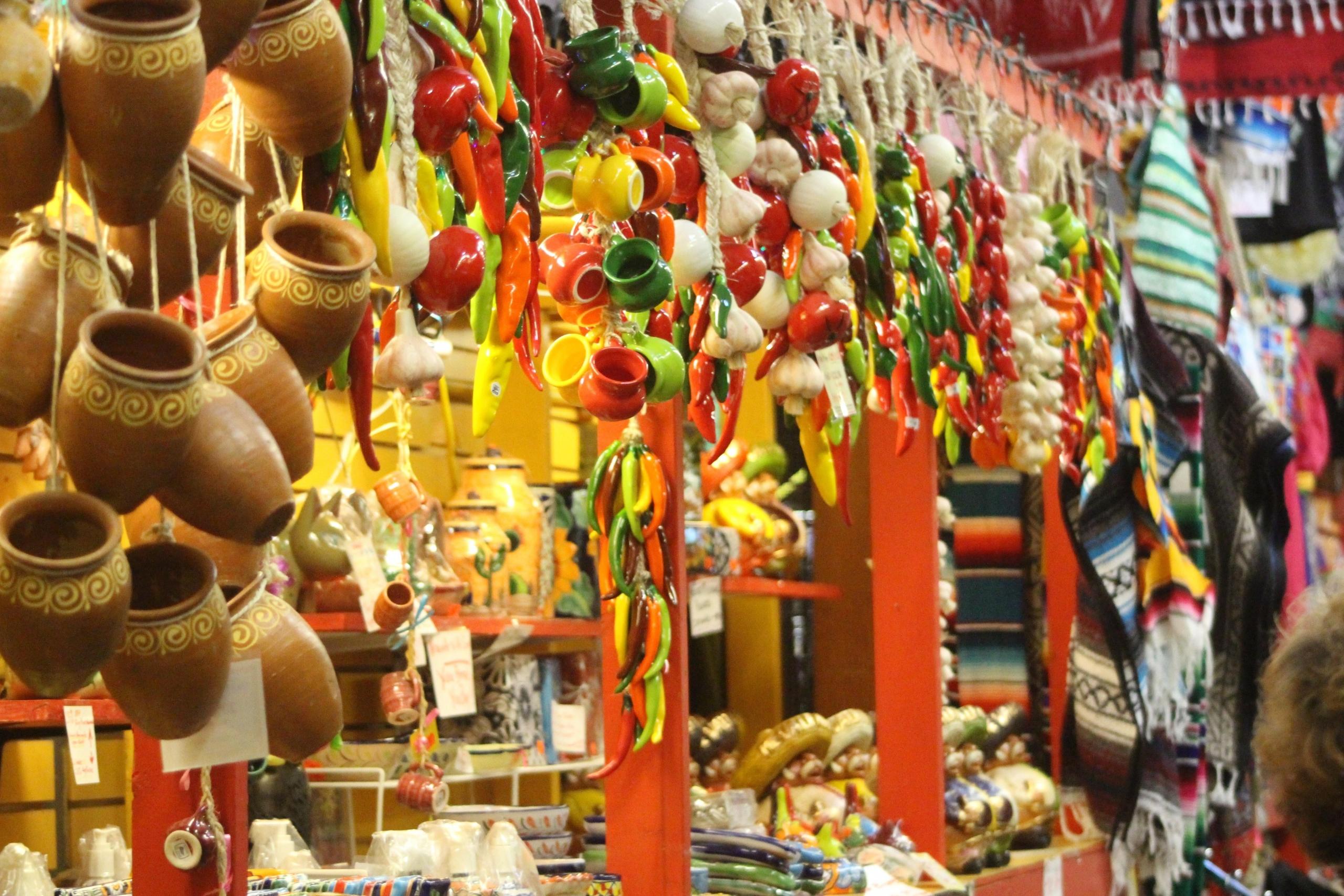 A view of a Mexican stall, an assortment of colored commodities are hanging, including garlic, peppers, pots, and small china mugs. There are also clothes hanging in the background. Mexican pop culture is as vibrant as the contents of this stall